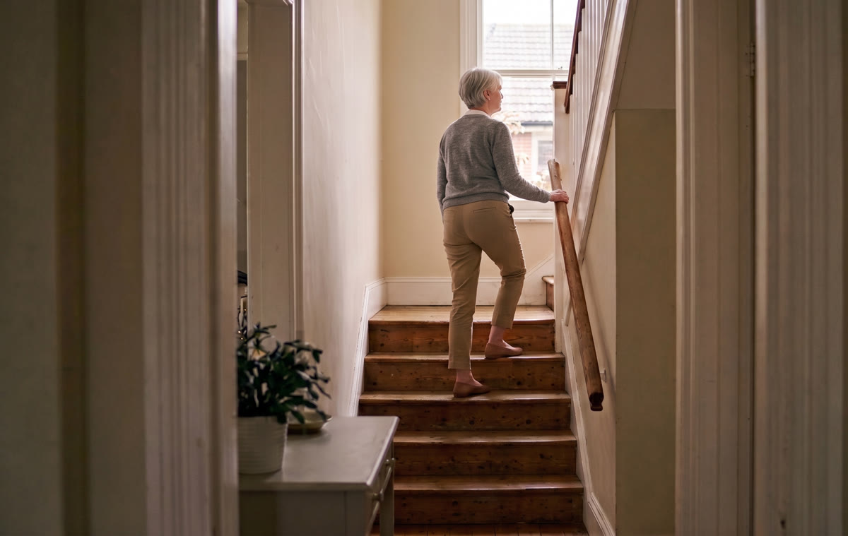 An older woman climbing the stairs at home, one hand resting on the handrail.